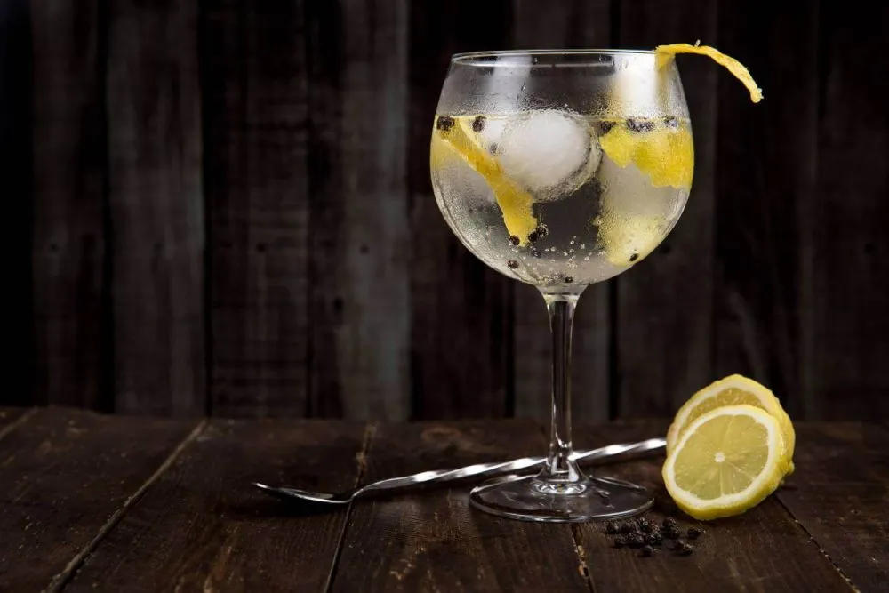 A full cocktail glass with lemon garnish sitting on a dark wooden table with a cocktail spoon and lemon slices, in front of a dark wooden board background.